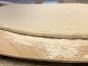 Close up image of pizza 1/8" thick pizza dough on a heavily floured, wood pizza peel.