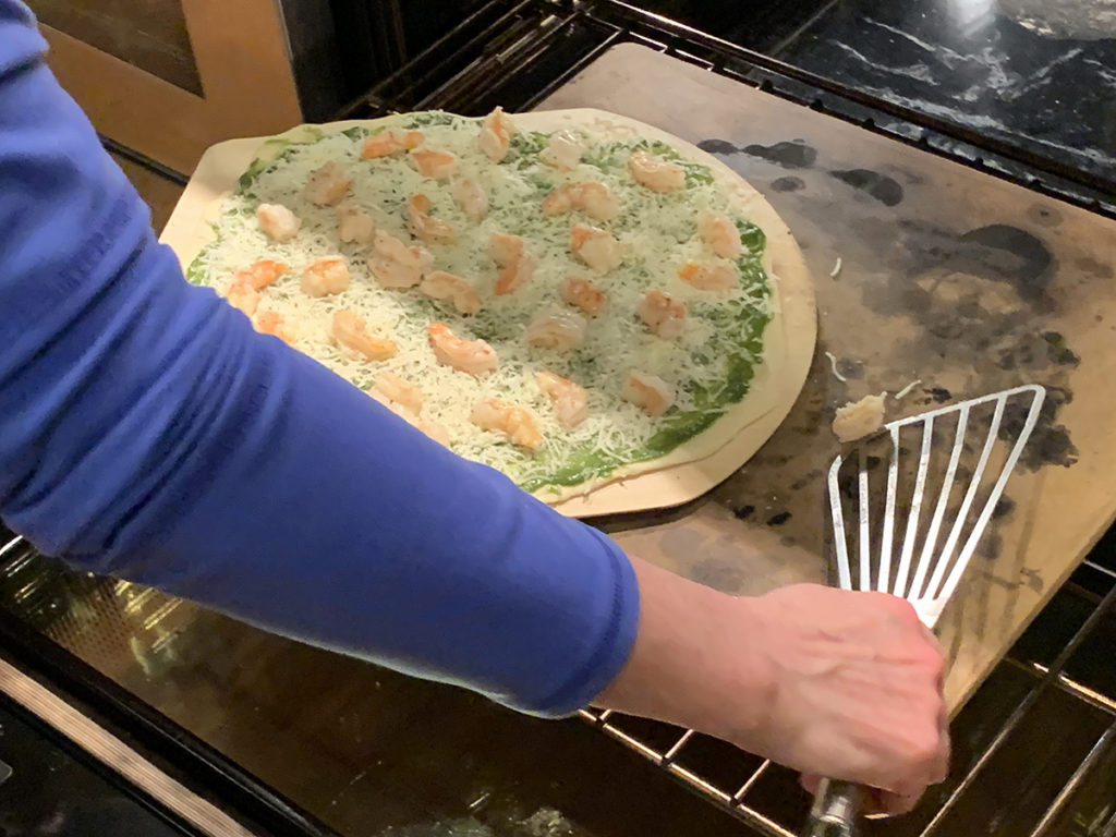 Pizza being transferred from a wood pizza peel to a pizza stone in an oven. There's a woman's arm scooping up a rogue shrimp that has fallen off the pizza with a metal spatula. 