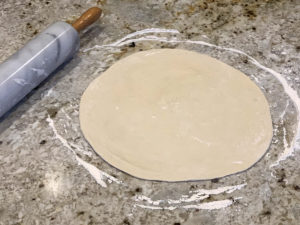 Round rolled out pizza dough on a floured granite surface with a marble rolling pin.