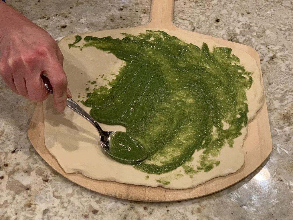 Female hand spreading bright green nettle pesto on a rolled out pizza dough with a metal spoon. Dough is on wooden pizza peel and that's on a granite surface. 