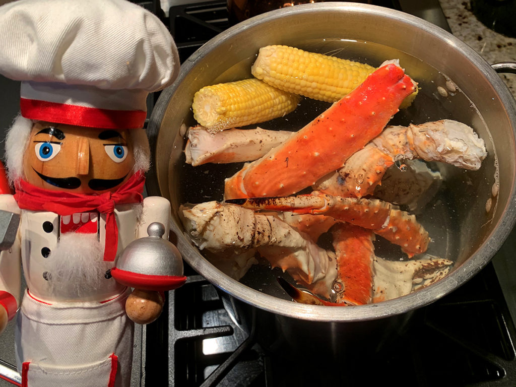 Alaskan King Crab legs and two ears of corn cooking in a large pot. There's a nutcracker who looks like a chef in the foreground. 