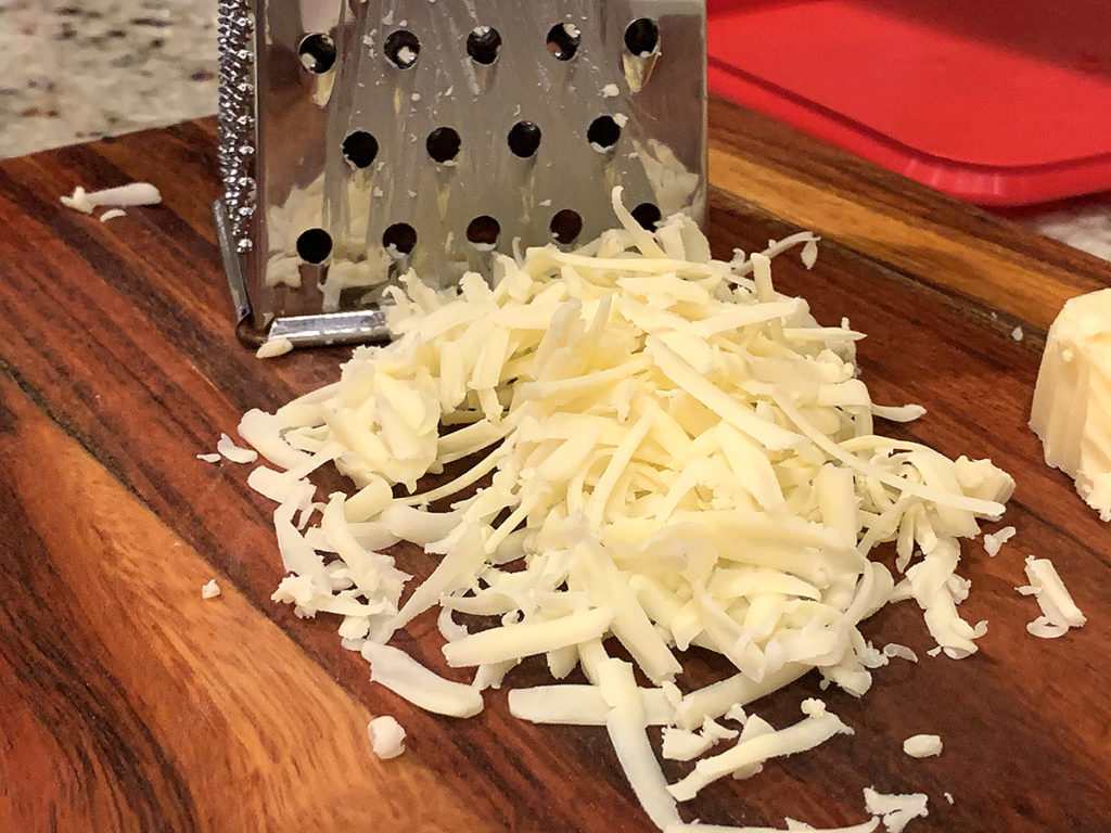 Sharp white cheddar cheese being grated with a box grater on a wood cutting board. 