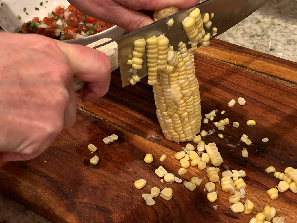 Hands using a chef knife to cut corn off of the cob. 