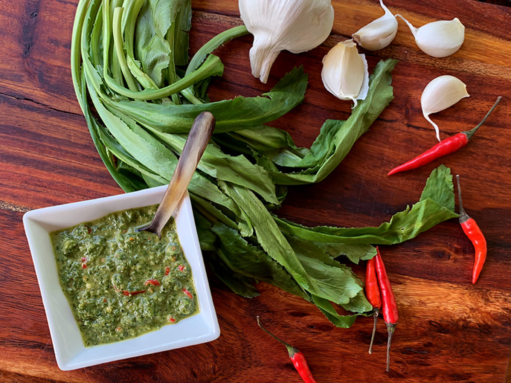 Culantro leaves, garlic cloves, whole red Thai chilies and a small white dish with a green sauce, all sitting on a pretty wood board.