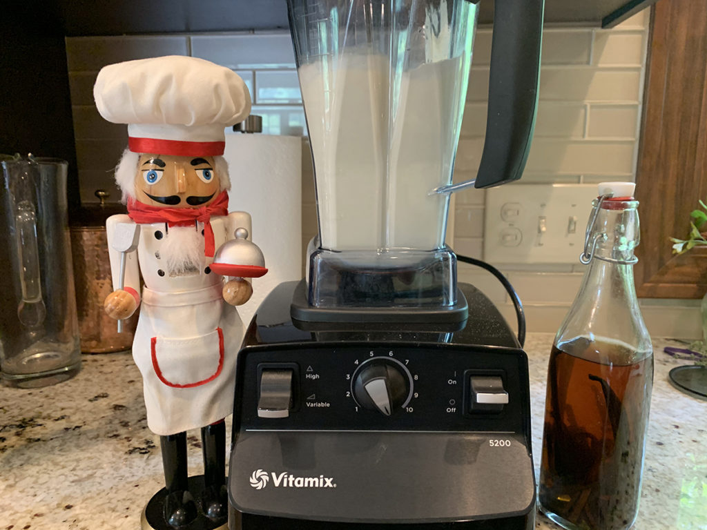 VitaMix blender with a pale yellow batter being blended. There's a nutcracker who looks like a chef to the left and a bottle of vanilla to the right.