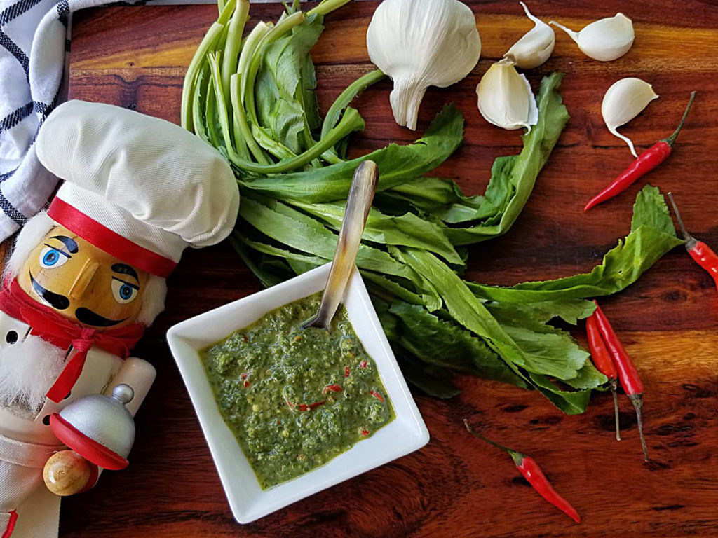 Culantro leaves, garlic cloves, whole red Thai chilies and a small white dish with a green sauce, all sitting on a pretty wood board. There's a nutcracker in the foreground who looks like a chef.