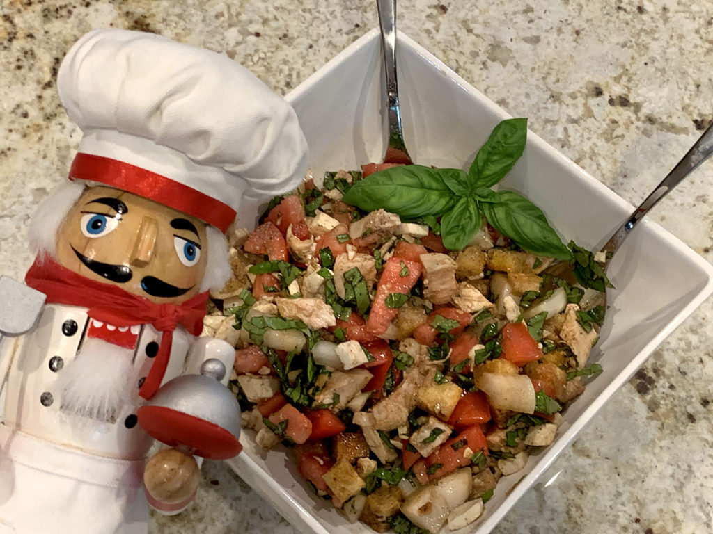 Chicken Panzanella Salad in a square white bowl with a fresh basil garnish and a nutcracker in the foreground who looks like a chef. 