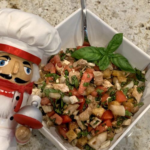 Chicken Panzanella Salad in a square white bowl with a fresh basil garnish and a nutcracker in the foreground who looks like a chef.