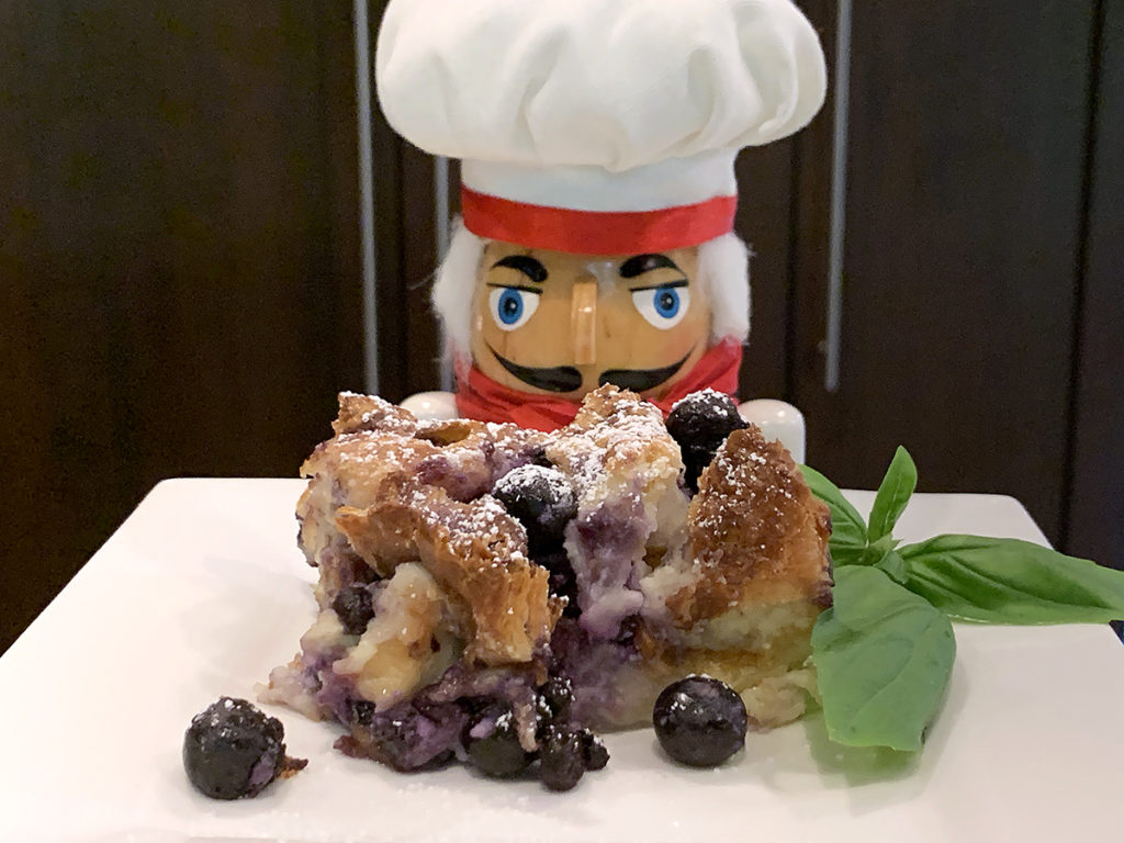 Blueberry bread pudding on a white plate with fresh basil garnish and a nutcracker who looks like a chef in the background
