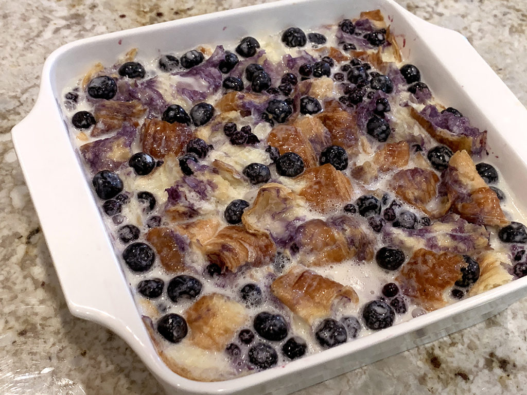 Batter soaking into blueberries and croissant pieces in a white square casserole dish ready to bake this blueberry & cream cheese croissant puff.