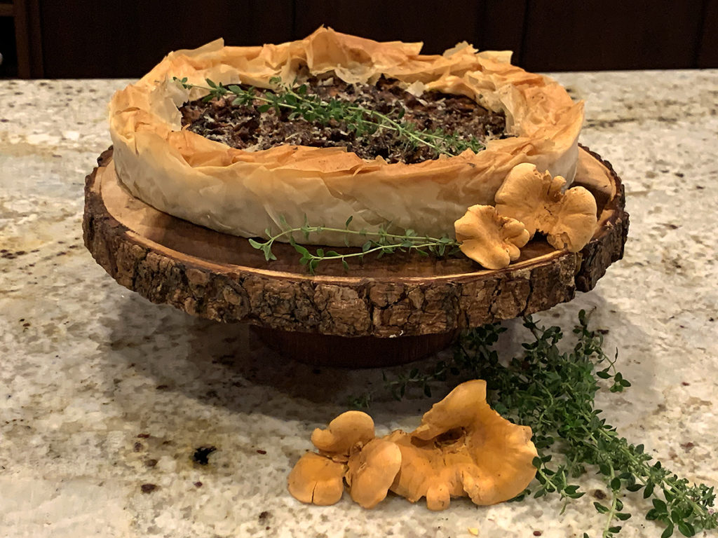 Rustic chanterelle tart with ruffled filo dough crust sitting on a round wood pedestal that has bark around the edge. There are a few raw, whole chanterelles and some fresh thyme on the board for garnish.
