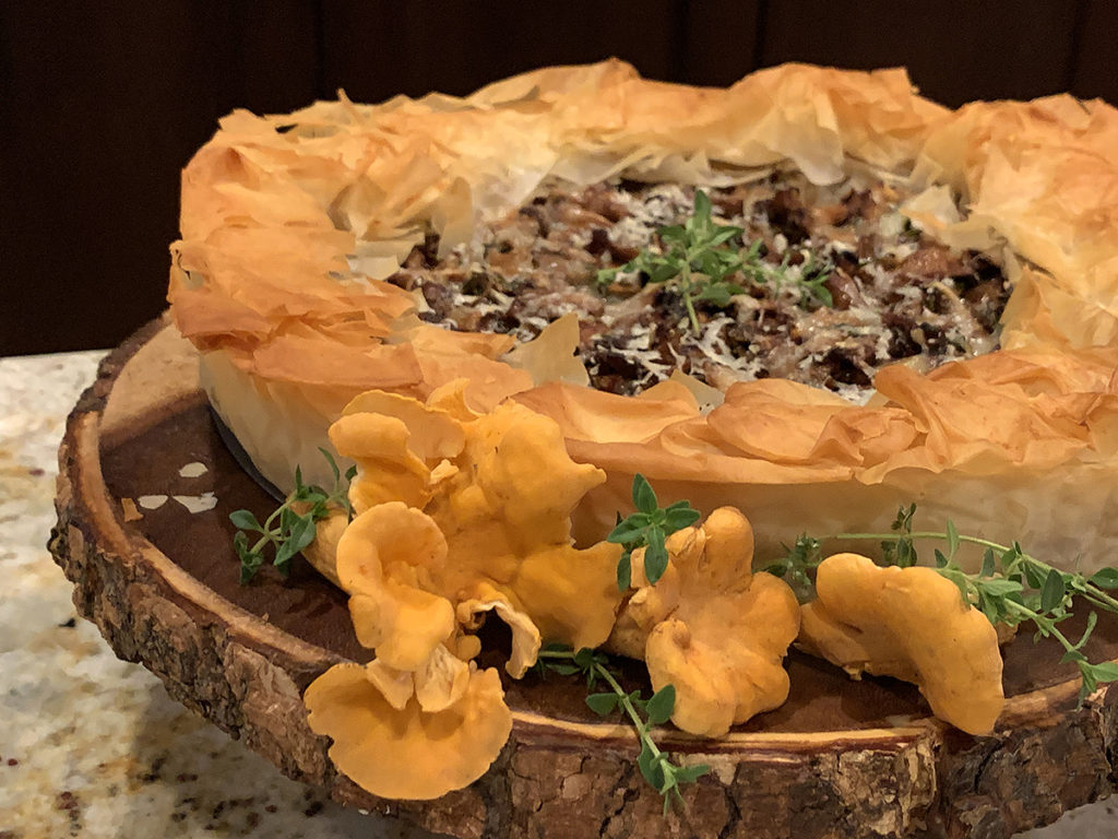 Rustic chanterelle tart with ruffled filo dough crust sitting on a round wood pedestal that has bark around the edge. There are a few raw, whole chanterelles and some fresh thyme on the board for garnish. 