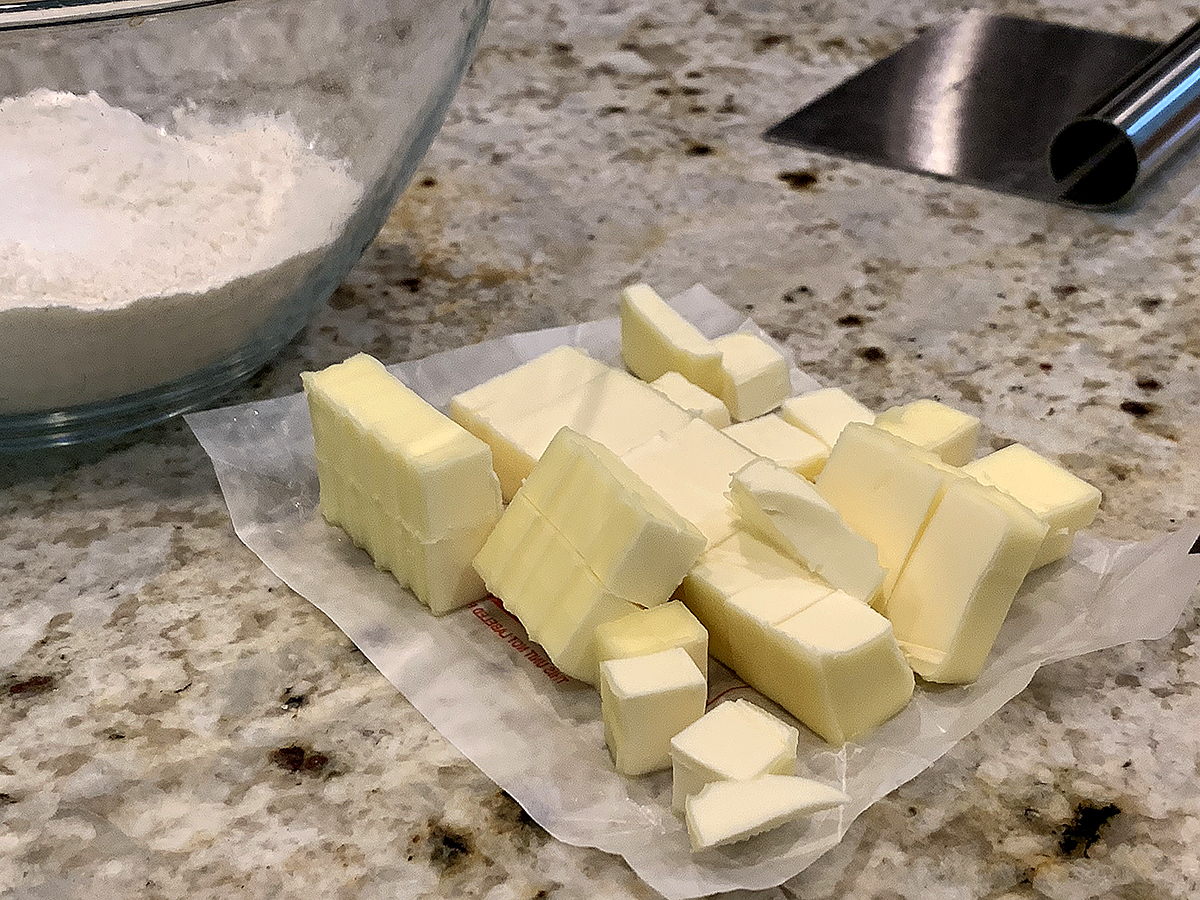 Butter cut into cubes sitting on the wax paper it comes in. There's a glass bowl with flour to the left and a steel bench scraper to the right. 