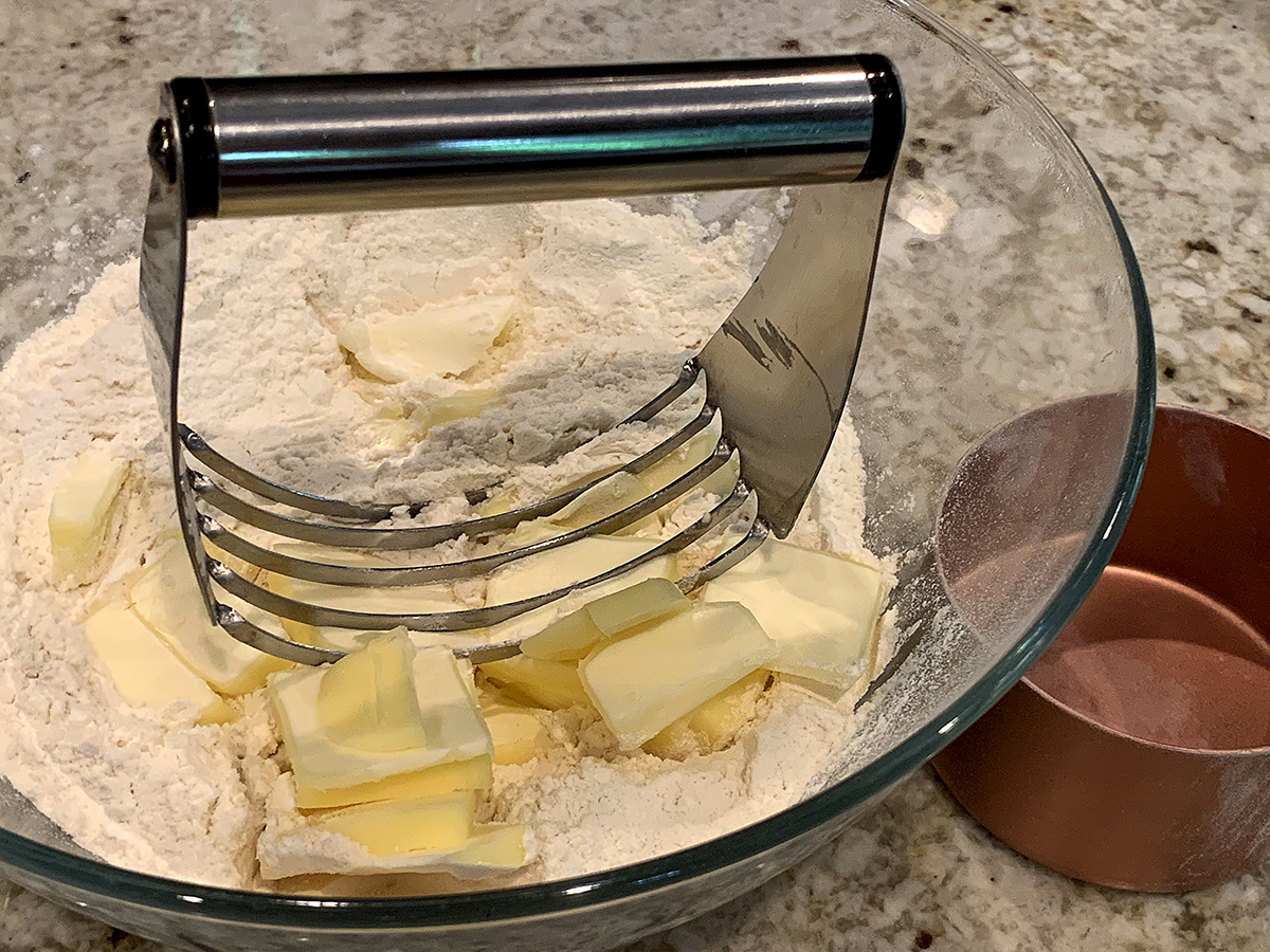 Cubes of butter and a pastry cutter on top of a pile of flour in a glass bowl.