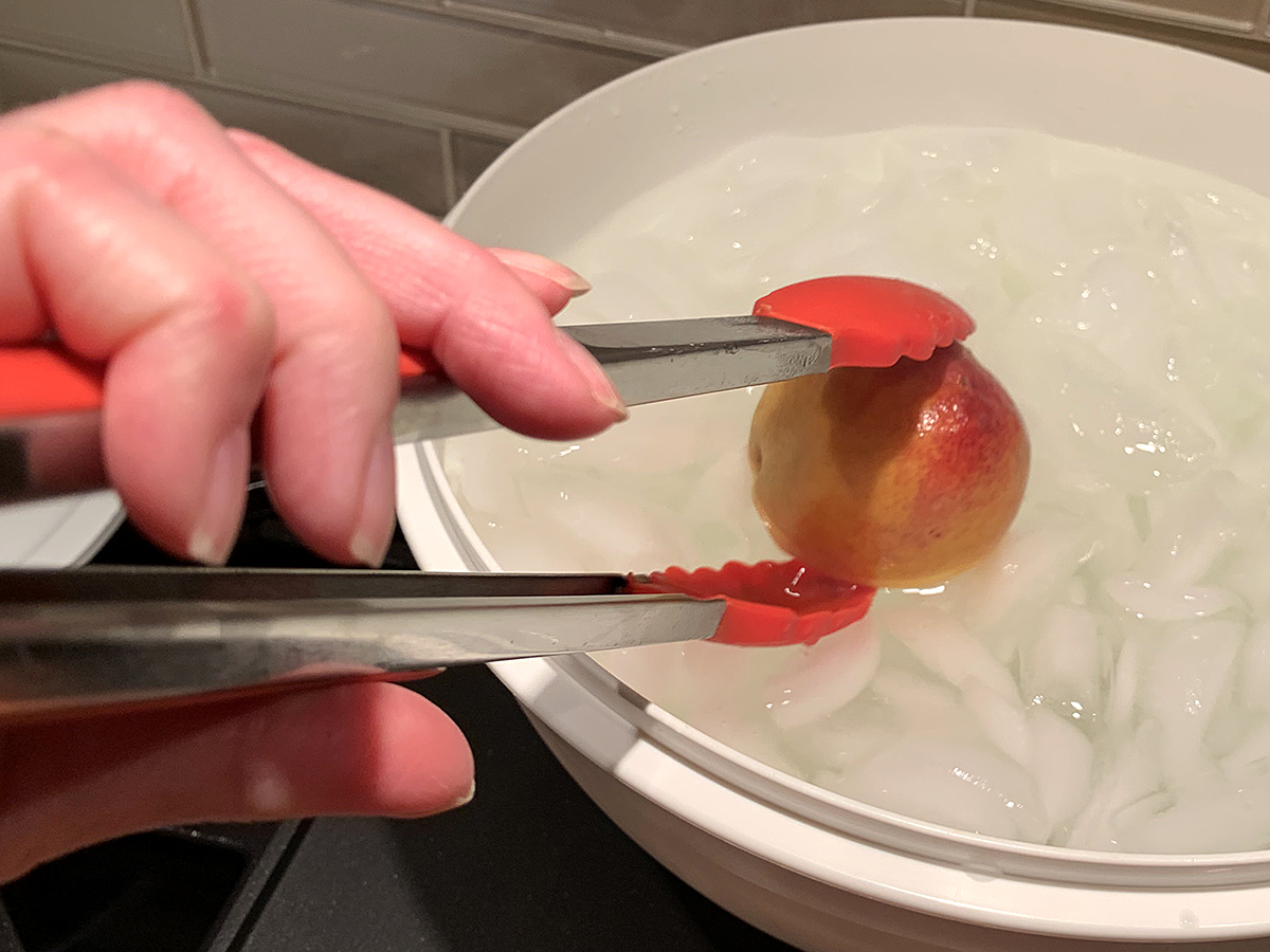 A large white bowl with boiling water, and a hand holding tongs that are lowering a peach into the ice water. 