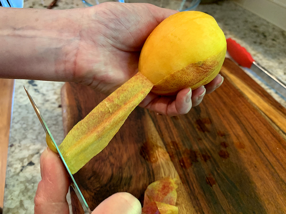 One hand holding a peach, while the other uses a paring knife to peel the skin away. 