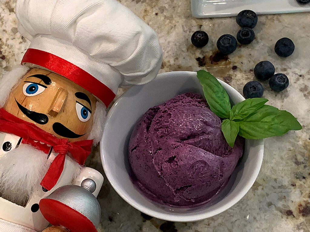 Scoops of blueberry ice cream in a small, white, round bowl and garnished with basil. Fresh blueberries in the background. There's a nutcracker who looks like a chef in the foreground.
