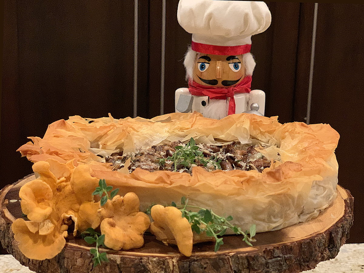 Rustic chanterelle tart with ruffled filo dough crust sitting on a round wood pedestal that has bark around the edge. There are a few raw, whole chanterelles and some fresh thyme on the board for garnish and a nutcracker who looks like a chef in the background.