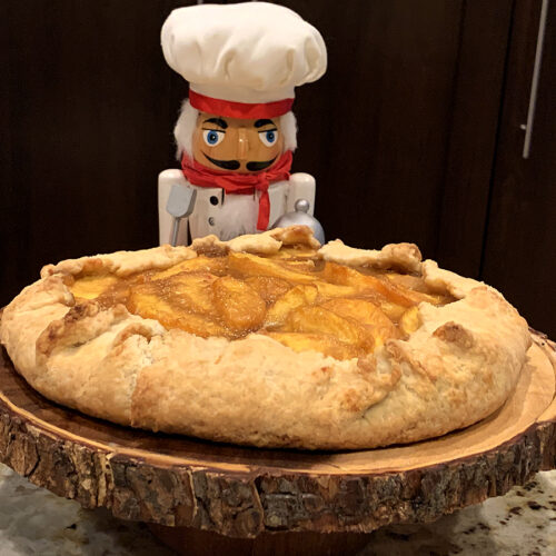 A rustic peach galette sitting on a round wood pedestal with a bark edge. There's nutcracker who looks like chef standing behind.