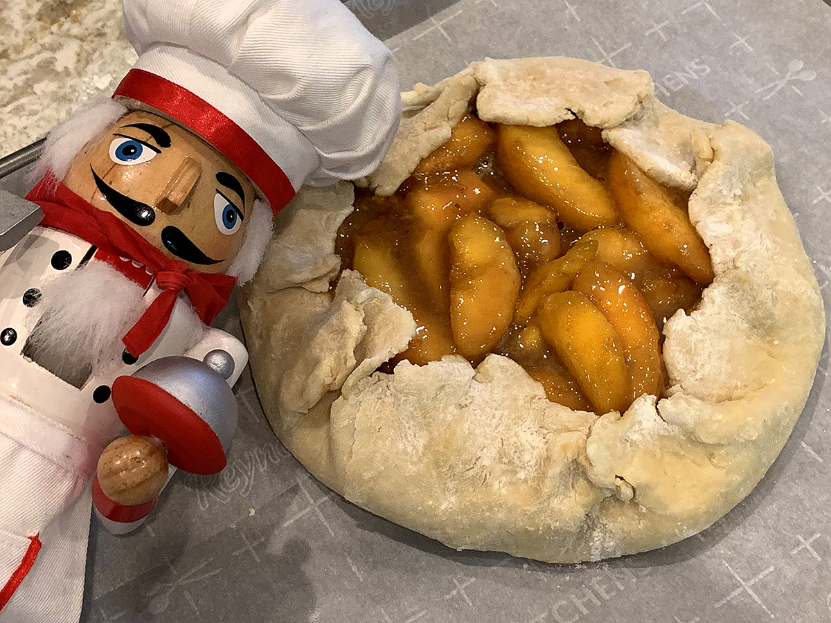 Unbaked peach galette with it's rustic crust on a parchment lined sheet pan. There's a nutcracker who looks like a chef in the foreground to the left. 