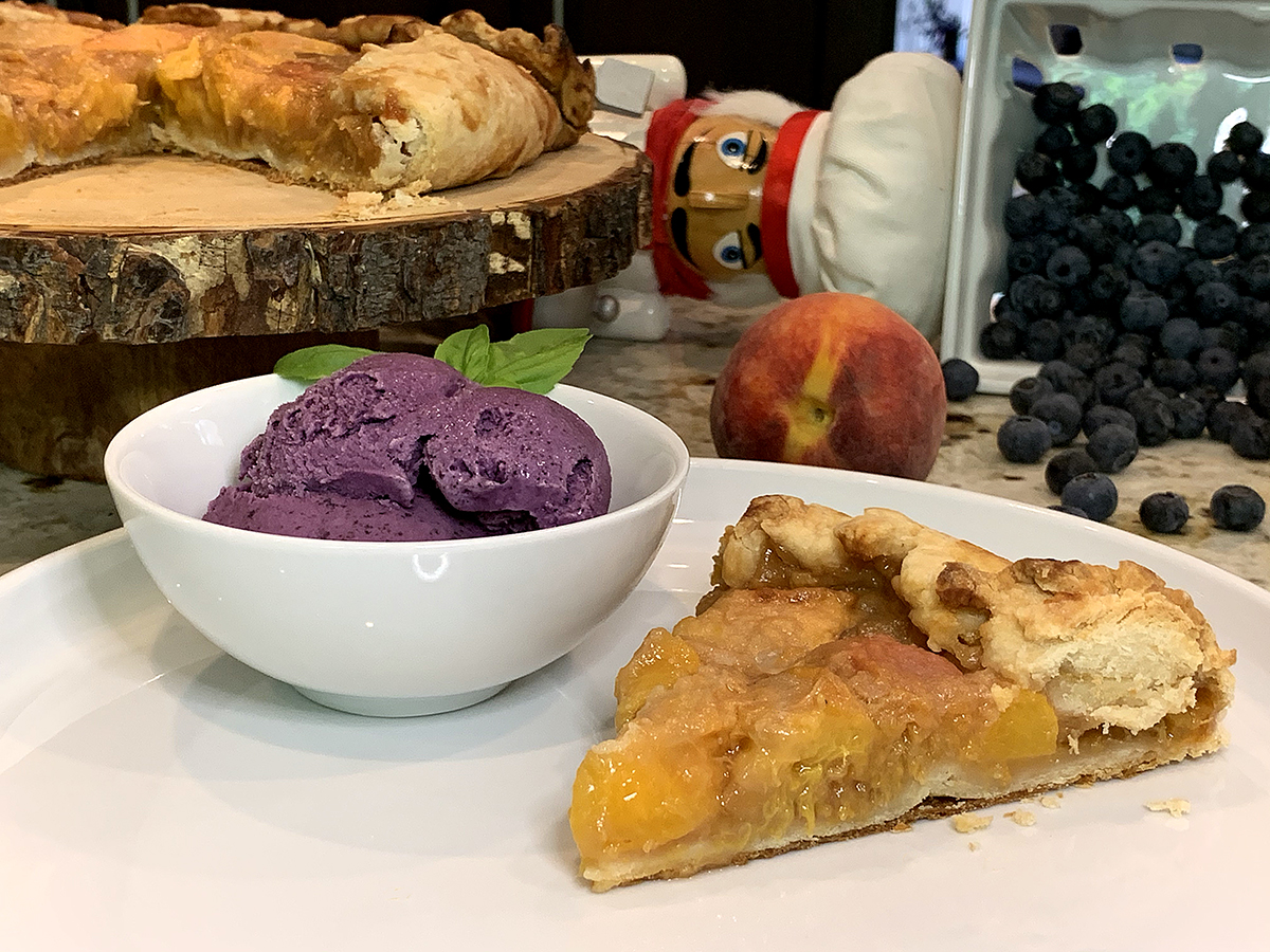 A small white bowl with Blueberry ice cream garnished with fresh basil, a slice of peach galette on a white plate. There's a nutcracker, fresh peaches and blueberries in the background.