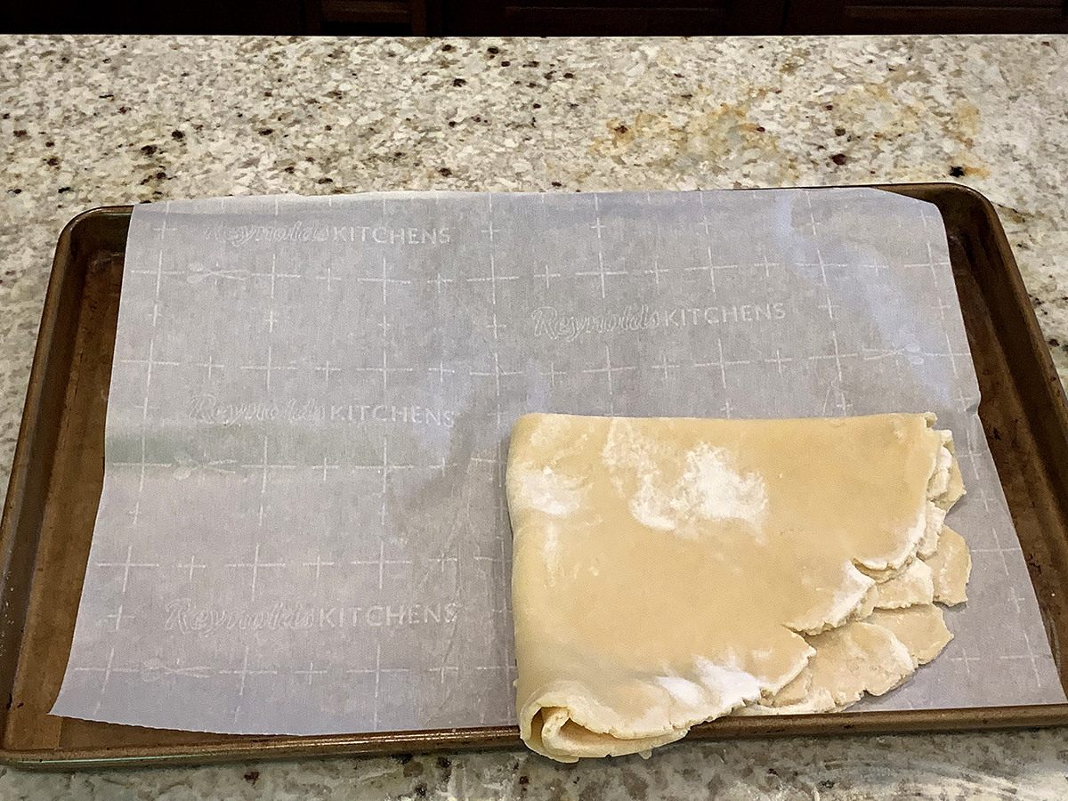 Rolled out pastry dough that has been folded into fourths and is sitting on the bottom right corner of a parchment lined sheet pan.