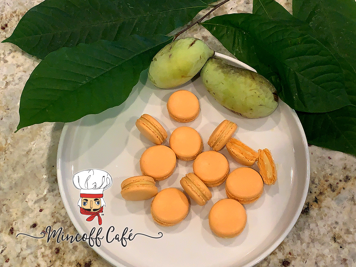 pale orange pawpaw french macarons on a round white plate with fresh pawpaw fruit and pawpaw leaves.