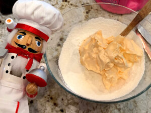 Pale orange meringue being mixed with almond flour and sugar in a clear bowl. There's a nutcracker who looks like a chef in the foreground.