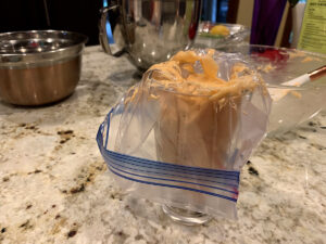 Pale orange macaron batter placed in a resealable bag that's being held by a pint glass.