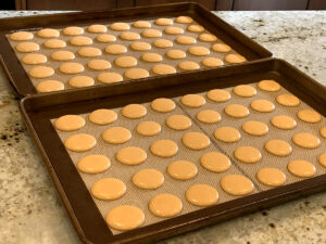 Two sheet pans with 20 round, orange disks of macaron batter.