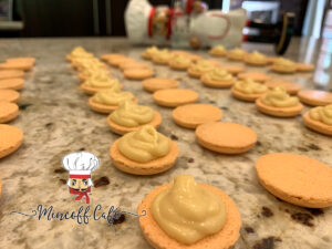 Pale orange round macaron shells face up, in rows on a granite counter top. Every other one has a pale yellow orange dollop of pawpaw white chocolate ganache on it. There's a blurry image of a nutcracker who looks like a chef in the background.