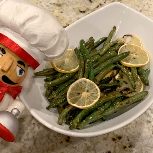 Roasted green beans with slices on lemon on top in a white bowl. There's a nutcracker who looks like a chef in the foreground.