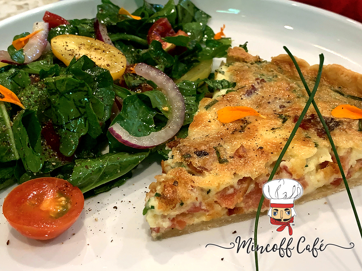 A wedge of bacon, chive and gruyere quiche wish a side salad, garnished with flower petals on a round white plate.