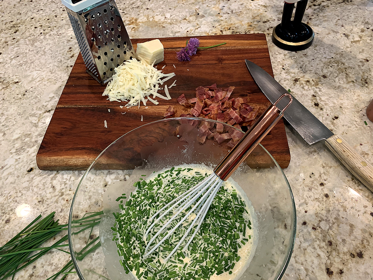 Clear bowl with beaten eggs and chives with a whisk in the bowl. Chopped bacon and shredded cheese on a wood board in the background. 
