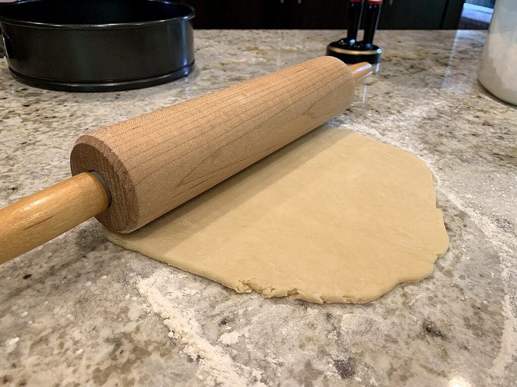 Pastry dough being rolled out with a wooden rolling pin on a granite countertop.