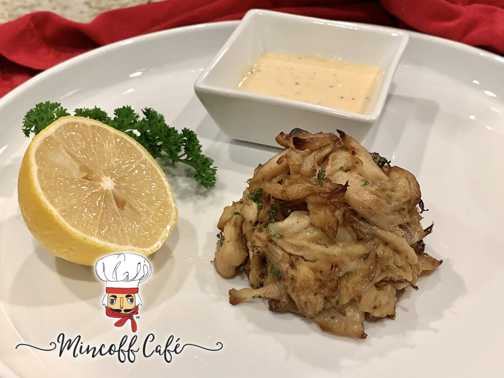 Crab cake mound on a round white plate with a square ramekin of pale yellow sauce, a lemon wedge and some parsley.