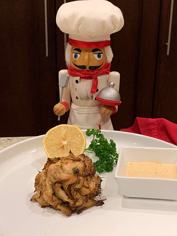 Crab cake mound on a round white plate with a square ramekin of pale yellow sauce, a lemon wedge and some parsley. There's a nutcracker in the background who looks like a chef.
