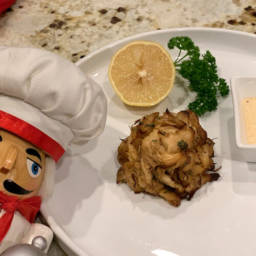 Crab cake mound on a round white plate with a square ramekin of pale yellow sauce, a lemon wedge and some parsley. There's a nutcracker in the foreground who looks like a chef.