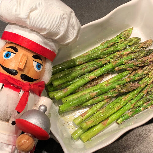 Bright green asparagus in a white rectangular dish. There's a nutcracker who looks like a chef in the foreground.