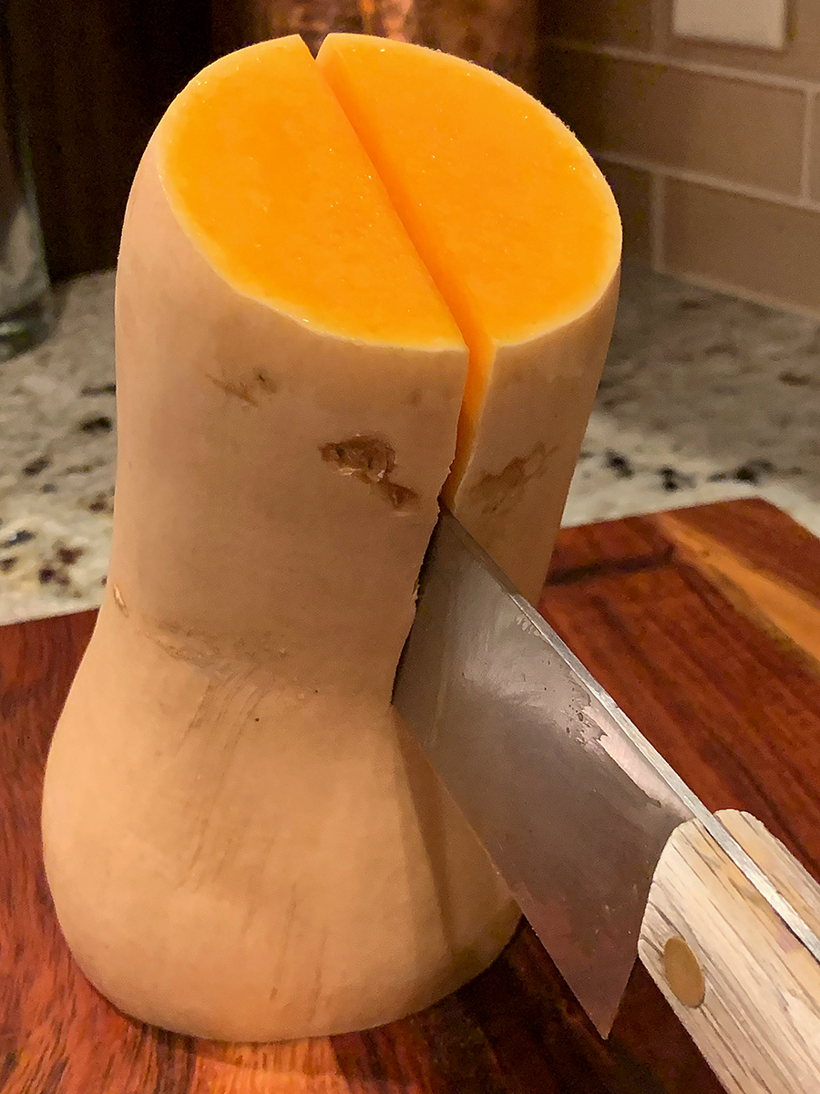 Butternut squash has it's ends cut off and is standing vertically on a wood board while being cut down the center.