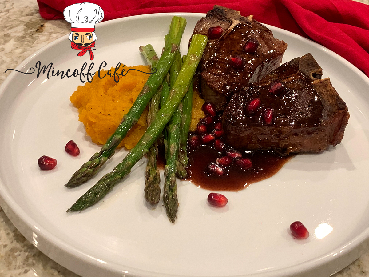 Two lamb chops with pomegranate wine sauce, fresh pomegranate seed, a few sprigs of asparagus and some mashed butternut squash on a round white plate.
