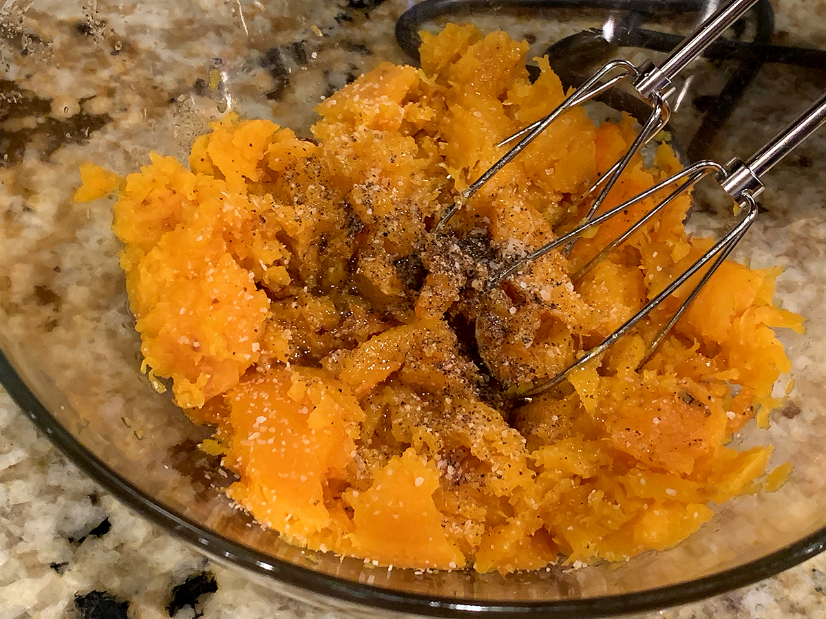 Amber colored butternut squash being mixed with a hand mixer in a clear bowl.