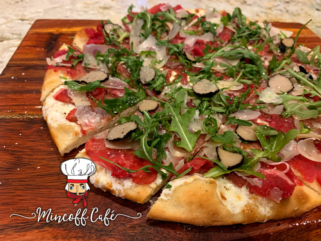 Beef carpaccio pizza with rare thin slices of beef, gooey cheese and arugula on a wood board.
