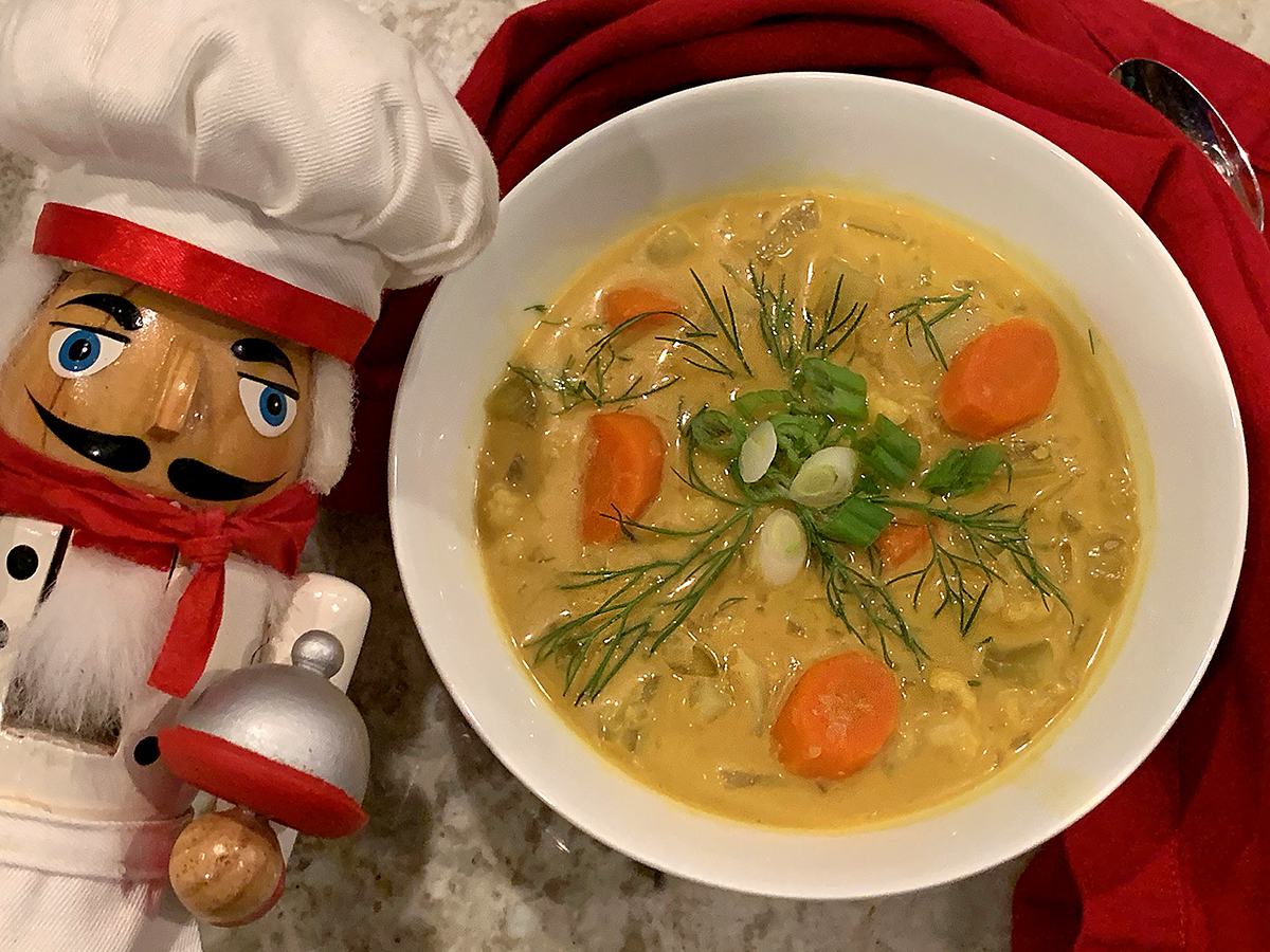 Pale yellow soup with Cauliflower, Fennel bulb and carrots in a round white bowl. Garnished with green onions, fennel fronds and chili oil. There's a nutcracker who looks like a chef in the foreground.