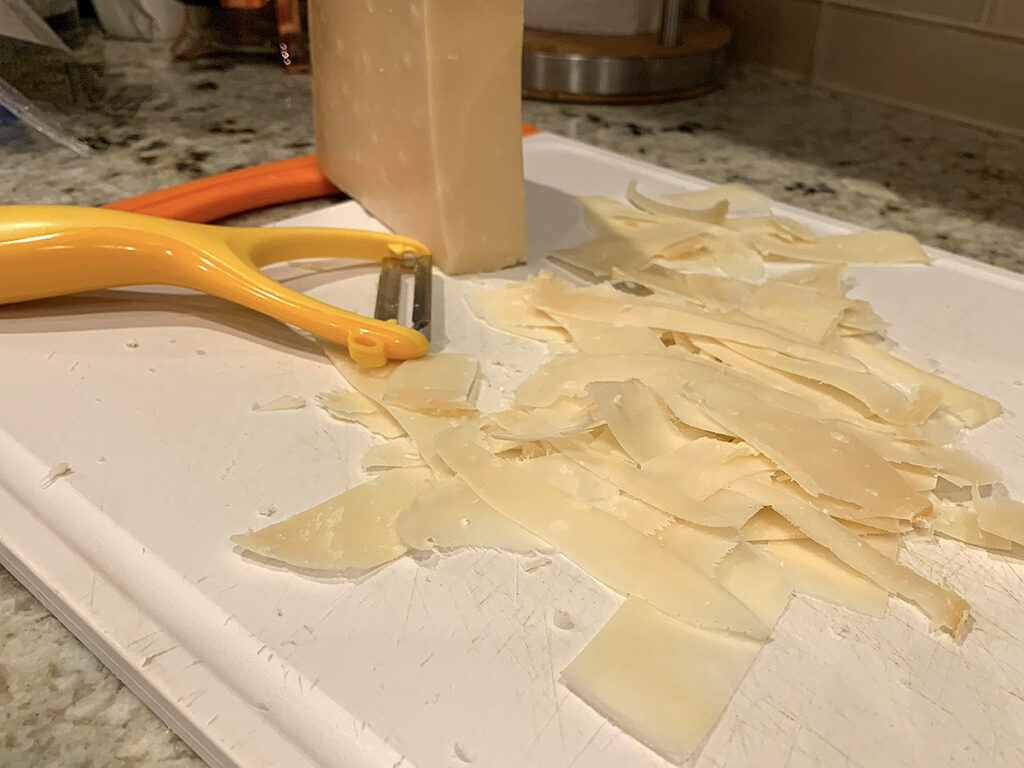 Parmesan cheese ribbons being sliced off block with vegetable peeler. 
