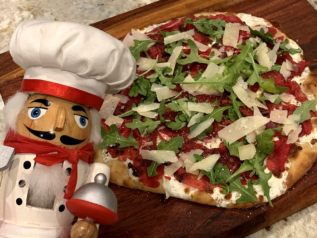 Beef carpaccio pizza with rare thin slices of beef, gooey cheese and arugula on a wood board. There's a nutcracker in the foreground who looks like a chef.