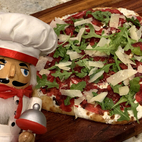 Beef carpaccio pizza with rare thin slices of beef, gooey cheese and arugula on a wood board. There's a nutcracker in the foreground who looks like a chef.