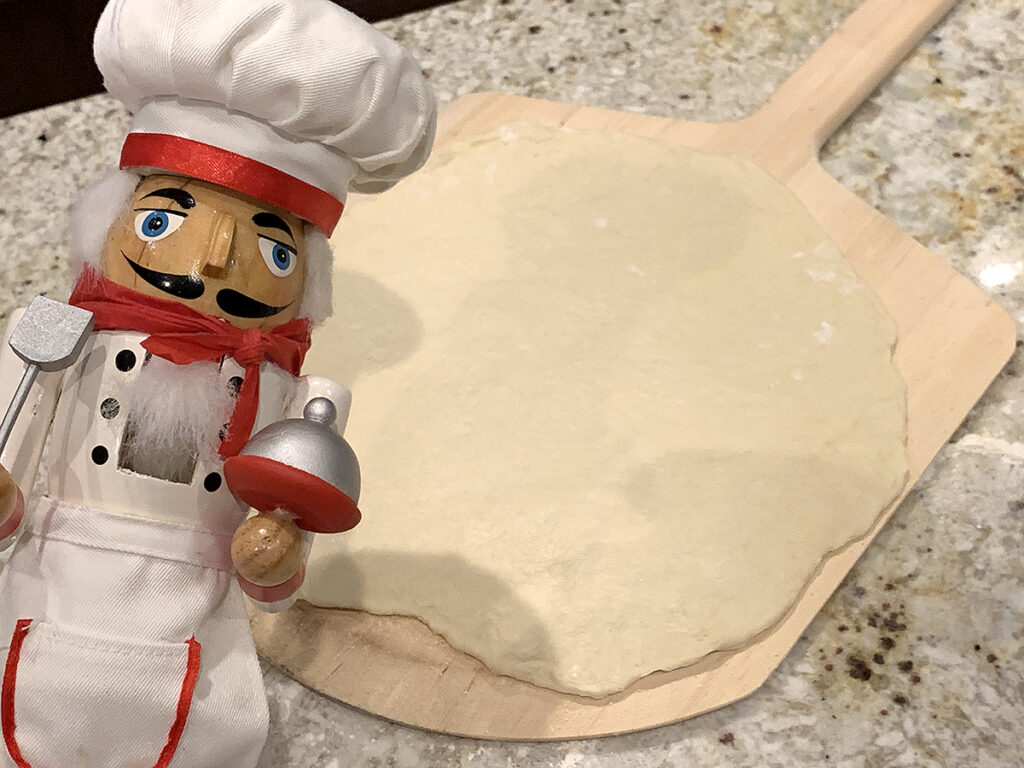 Homemade rolled out pizza dough laying on a wood pizza peel. There's a nutcracker in the foreground who looks like a chef.