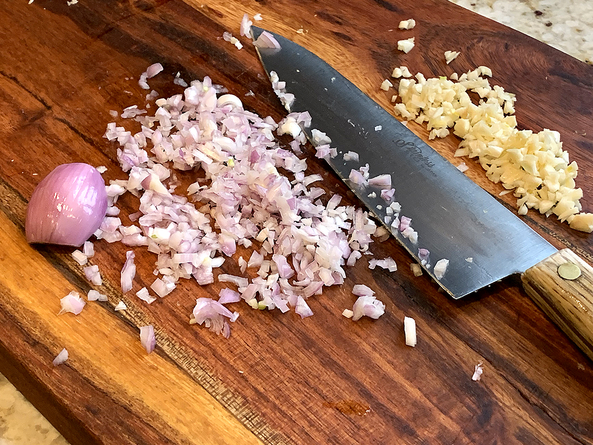 Minced shallot and garlic on a wood cutting board with a chef knife.