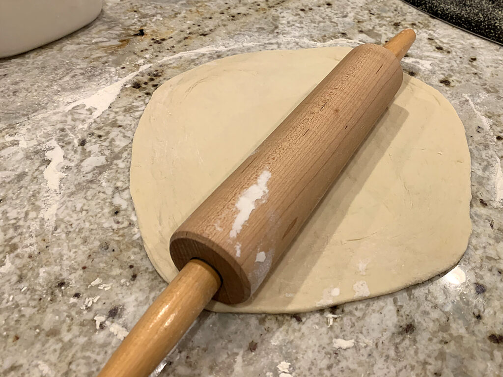 Round, rolled out pizza dough on a lightly floured granite countertop with a wood rolling pin on top.